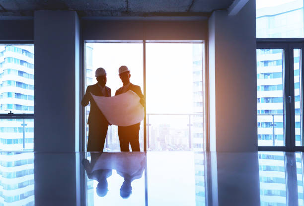 Engineer and businessman discussing project with big plan in empty office against window